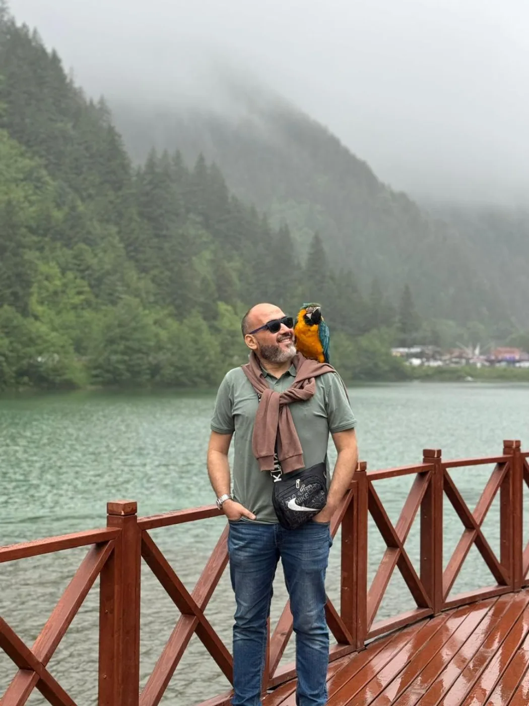 Dr. Abu-Sheikha on a wooden pier by a misty green lake, smiling at a macaw perched on his shoulder